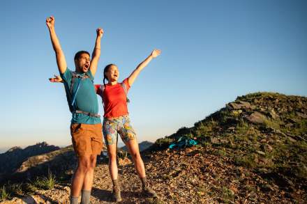 Man and Woman wearing hiking gear with their arms up and looking excited to reach the top of a mountain
