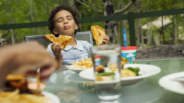 Kid with a very satisfied look on his face holding a sandwich. He is on a patio dining area with trees in the background