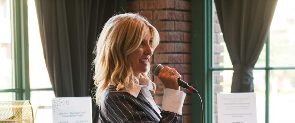 Kim Stowe speaking into a microphone standing in front of two tall tables with white tablecloths on them