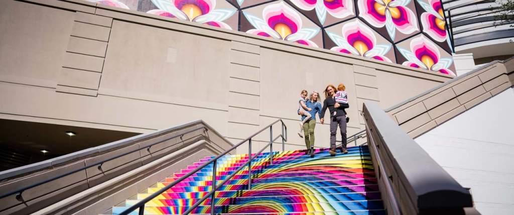 Family on stairs that are painted in a rainbow swirl with cherry blossoms on the wall behind them