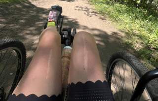 Wide natural trail with trees and grasses on each side. A recumbent quad cycle is biking down the path.