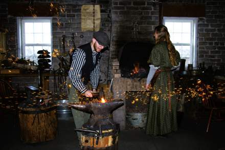Blacksmith striking a hot iron on the anvil and making sparks fly. A fire and a female blacksmith in period dress are in the background