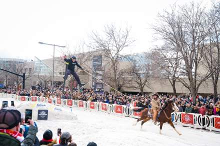 A crowd of people on both sides of a city street that is covered with snow and has a ski jump made from snow. In the center is a skier in the air after going off the jump being towed by a horse in traditional ski-joring.