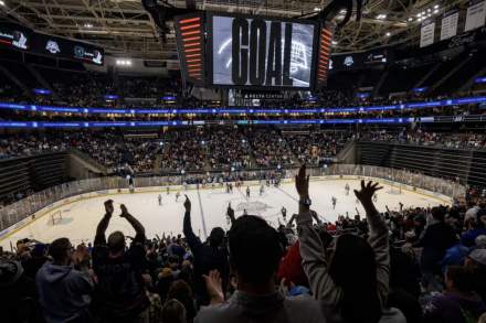 Hockey arena with fans cheering. scoreboard that reads GOAL, while players are on the ice sheet.