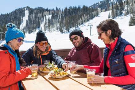 4 people sitting at an outdoor table with food, solitude mountain with snow and a lift in the background